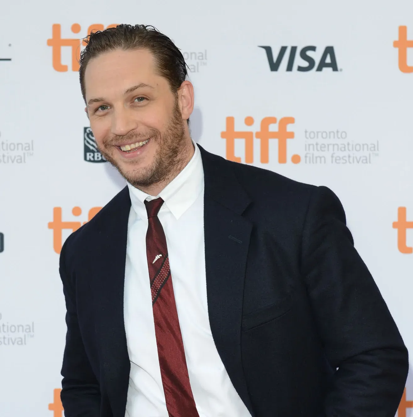 Tom Hardy smiling with a visible gap where a tooth appears to be missing, likely part of a film role or temporary dental effect, captured in a candid moment.